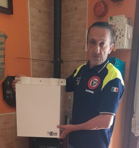 Sergio Attolini, vicepresidente dell'unità di Protezione Civile Radio i Due Falchi, in uniforme, con il filtro ProOzone donato, in piedi in una stanza con pareti arancioni.